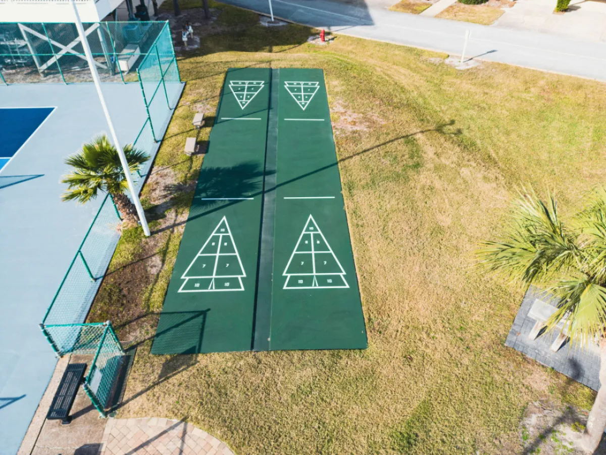 A bustling outdoor scene featuring shuffleboard courts, surrounding greenery, and tennis courts, where people can play games, socialize, or simply enjoy the sunny ambiance. Benches and communal sports areas set the tone for friendly competition and group activity.
