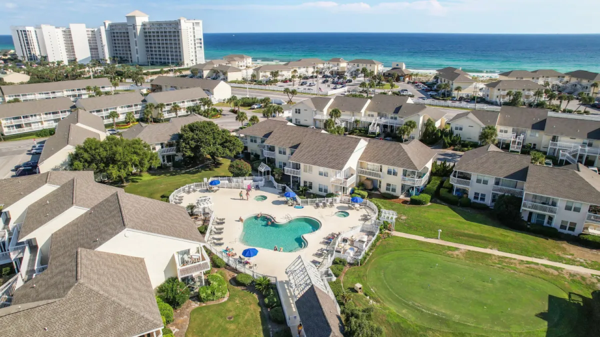 A lively swimming pool surrounded by lush greenery, with the beach visible in the background and residential buildings beyond. The pool area has space for groups to gather, sunbathe, and enjoy the resort's amenities in a social, outdoor setting.
