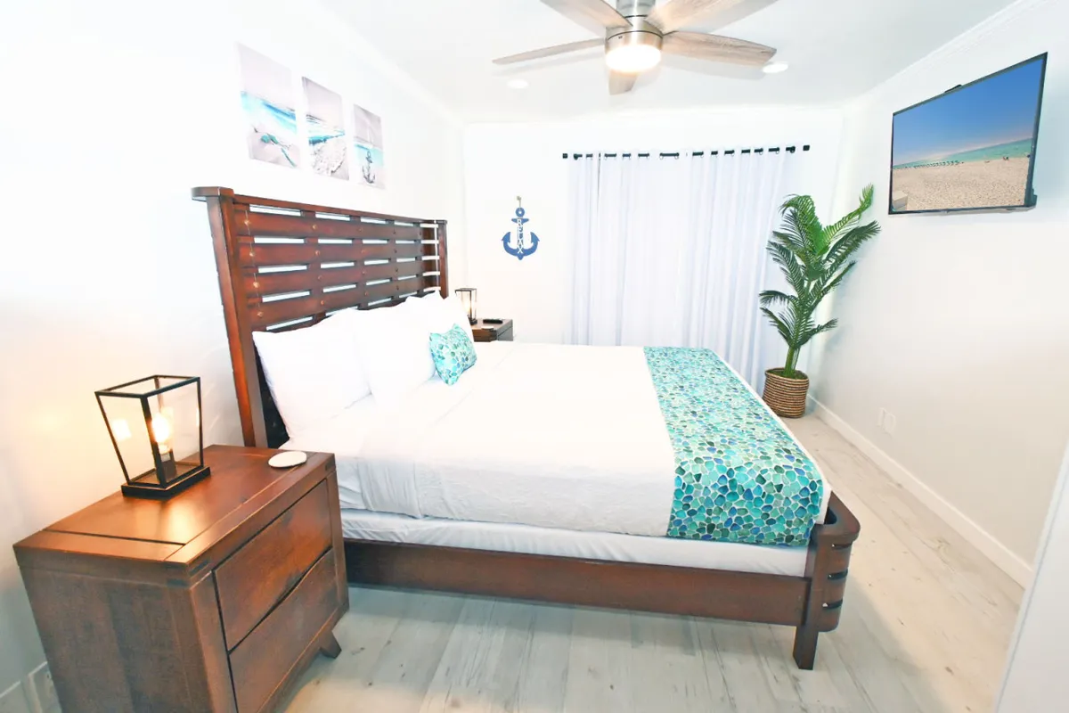 A tranquil bedroom with a bed decorated in soft, neutral bedding, a nightstand with a lamp, a tropical plant, beach-themed wall art, and a television opposite the bed. The space is filled with natural light, creating a restful and inviting atmosphere.