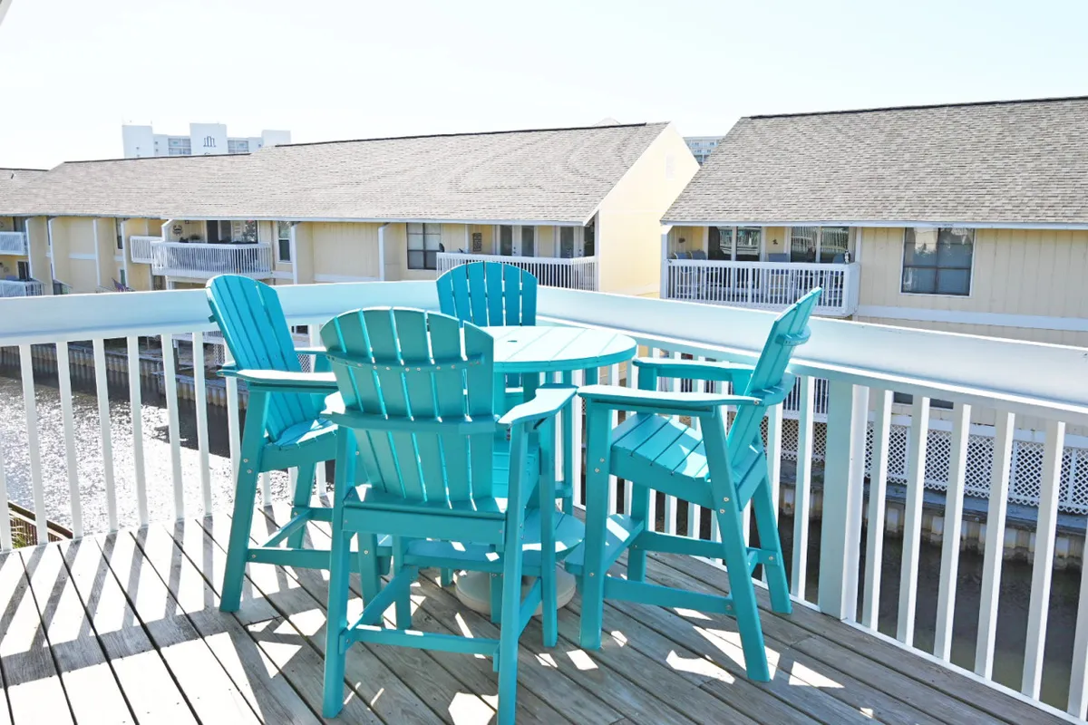 This image features a cheerful wooden deck with turquoise patio furniture overlooking a tranquil water view. The setting is inviting, suggesting a space perfect for outdoor meals, gatherings, or simply relaxing with a book in the sun. The light suggests either sunrise or sunset, enhancing the atmosphere of the deck as a place to unwind and enjoy nature.