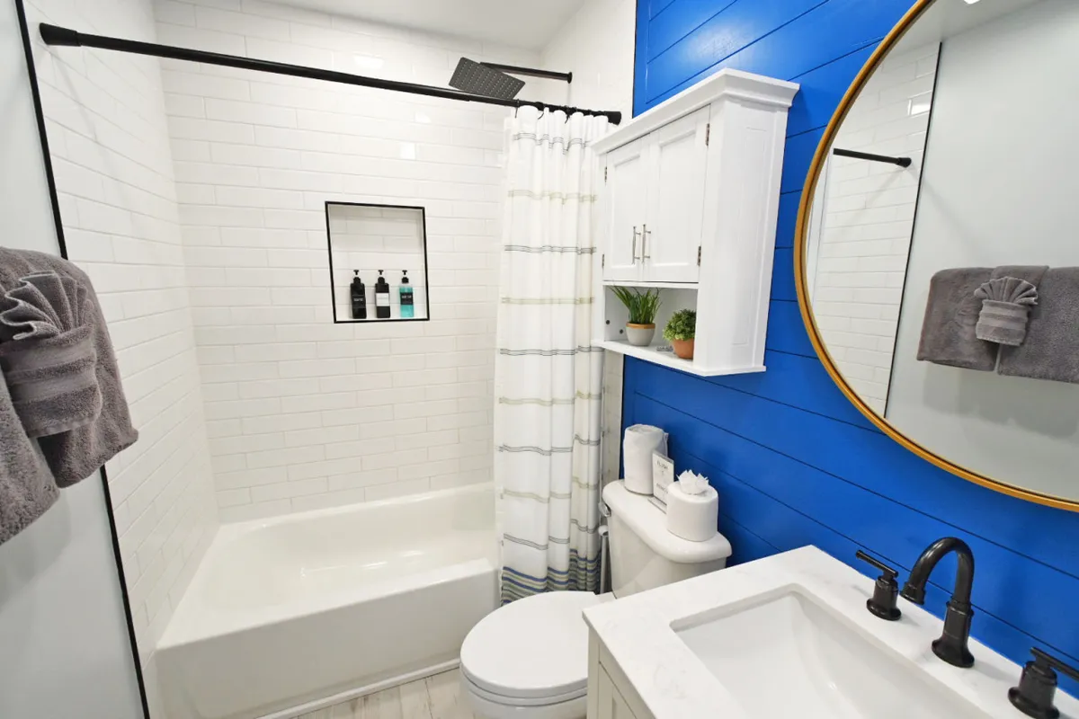 A bathroom interior with a blue accent wall, a circular mirror, and a visible shower area. The space looks fresh and inviting, emphasizing cleanliness and spa-like relaxation.