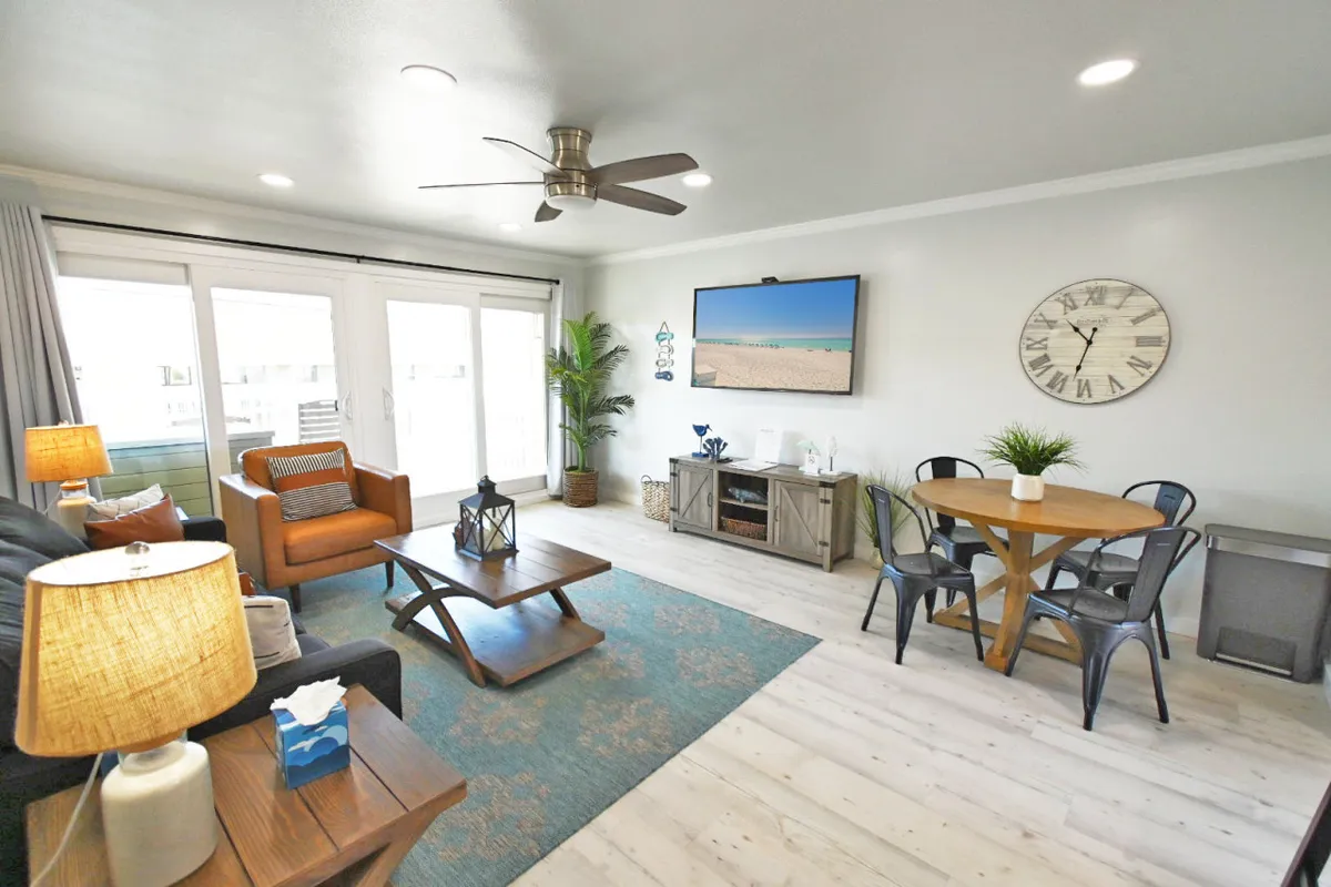 A bright, stylish living area featuring a comfortable sofa, a modern dining table for four, a large TV perfect for movie nights, and a sunlit window with gauzy curtains that fills the space with natural light. The inviting setup suggests a cozy spot for relaxing, sharing meals, or unwinding as a family.