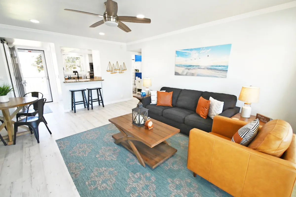 A cozy living space featuring a plush sofa, coffee table, wall artwork, and a view into the adjoining dining area and kitchen bar. The ambiance suggests a space ideal for unwinding, socializing, and enjoying group activities, with glimpses of sunset views through the windows.