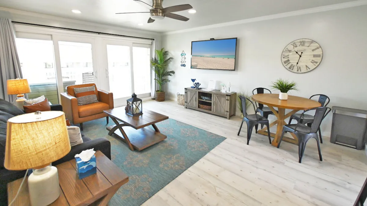 The image showcases a bright and inviting living space with a comfy sofa and armchair arranged around a mounted flat-screen TV, a stylish round dining table with modern metal chairs, and expansive windows/doors that open to a private balcony overlooking the tranquil canal. Sunlight fills the room, creating a warm, relaxing atmosphere ideal for unwinding after a day at the beach or enjoying a peaceful morning coffee with waterfront views.