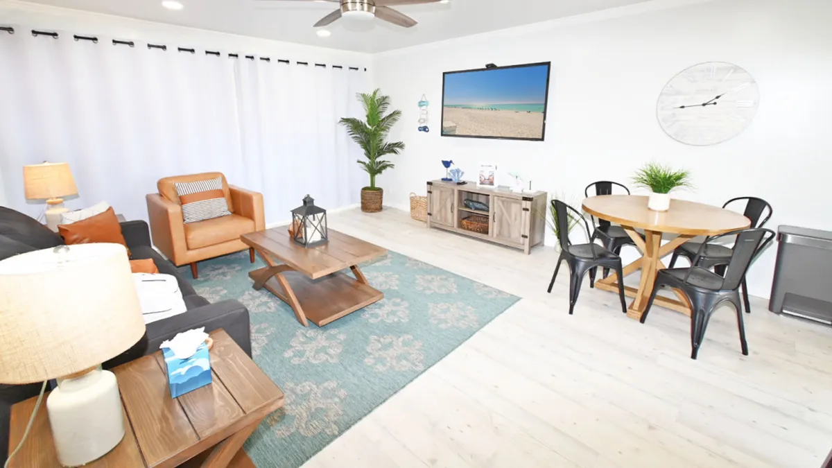 A cozy, light-filled living room featuring a dark gray sofa with orange and white pillows, a wooden coffee table with a lantern centerpiece, a flat-screen TV, and a round wooden dining table with black metal chairs. Large windows bring in natural light, and the decor is inviting and coastal-inspired.