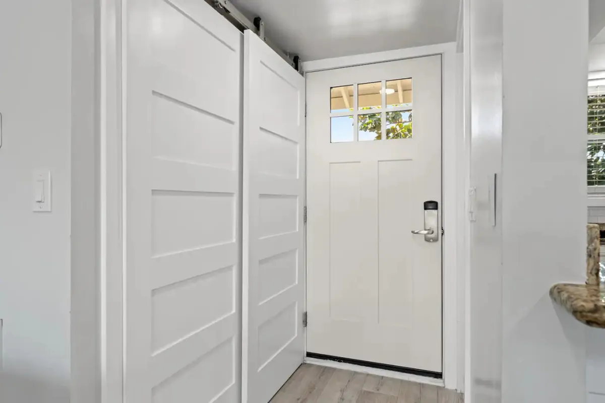 This interior image features a white front door with window panes, sliding barn-style closet doors, and part of a granite countertop. The light wood-style flooring and overall brightness create a welcoming, organized entryway. The closet area is visible, hinting at storage for beach towels and sports gear, tying directly to the section's mention of a well-stocked closet for guest convenience.