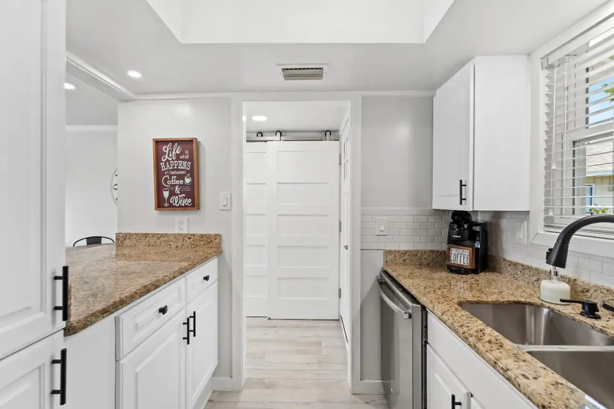 A bright, inviting kitchen area featuring granite countertops, white cabinetry with black handles, a stainless steel dishwasher, and a coffee maker on the counter. The space is organized and ready for meal preparation or morning routines.