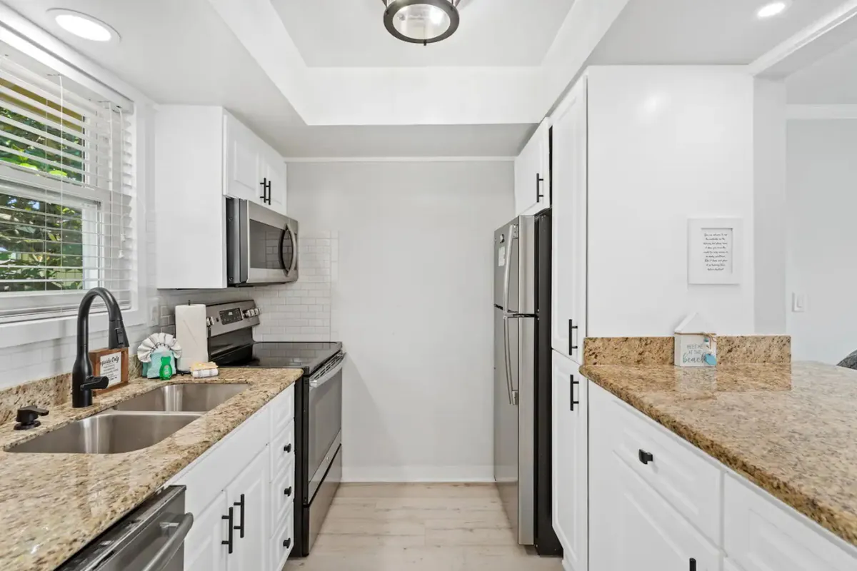 A modern kitchen with granite countertops, full-size stainless steel appliances, white cabinetry, and a kitchen sink with a black faucet. The space is well-lit, with ample counter space ideal for meal prep and casual socializing around the kitchen bar.