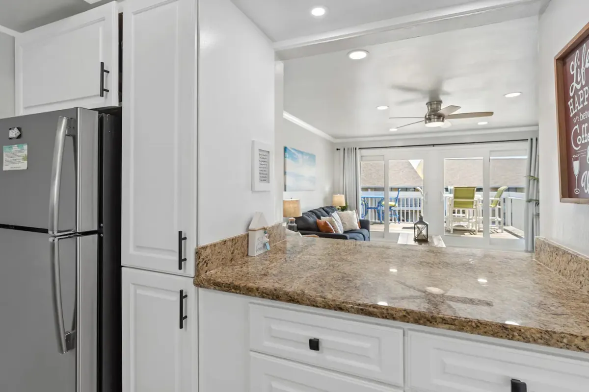 A bright, open-concept kitchen with granite countertops, white cabinetry, and modern black handles. The living room seating area and sliding glass doors to the balcony are visible in the background, highlighting the seamless flow between spaces.