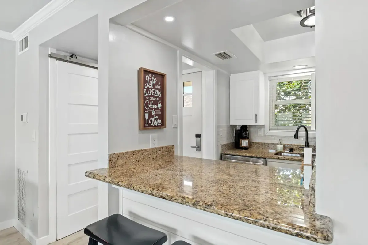 A welcoming kitchen bar area with a granite countertop, two stools, white cabinetry, a coffee maker, and a decorative wall sign reading 'Life happens between Coffee & Wine.' The setup is perfect for casual breakfasts or sharing stories over coffee.