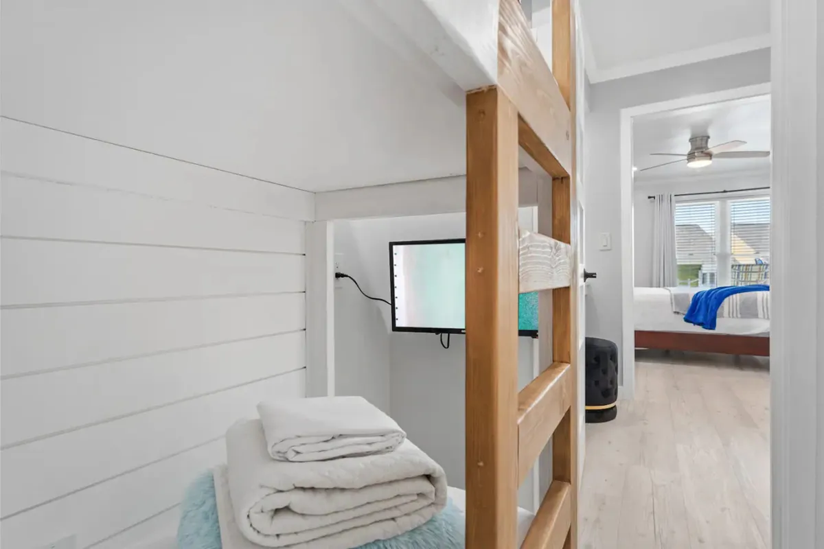 A bright bonus bunk room with wooden bunk beds and a sturdy ladder, a mounted flat-screen TV, neatly stacked white towels, and a partial view into the adjacent king bedroom. The room is tidy, cheerful, and clearly designed for comfort and functionality, ideal for kids or extra guests.