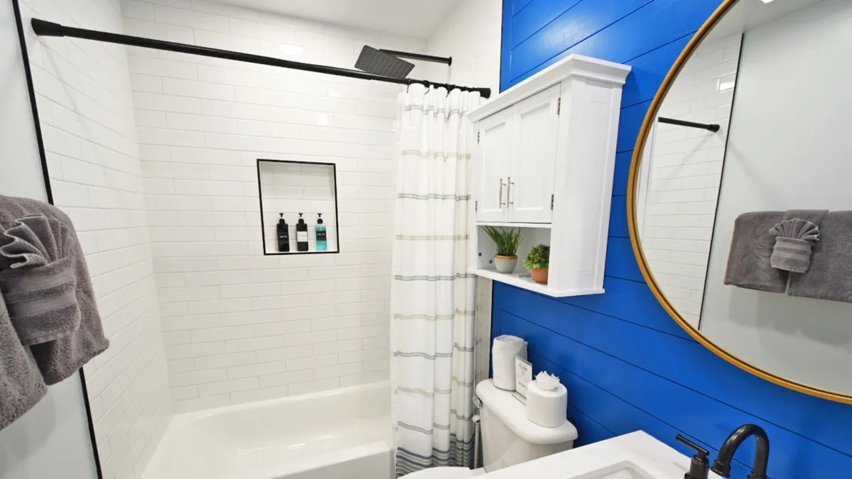 A modern bathroom with a white bathtub, built-in shower niche, black rain showerhead, large round mirror with a golden frame, blue shiplap accent wall, and a white cabinet adorned with small green plants. The space feels fresh, clean, and thoughtfully designed for relaxation or rejuvenation.