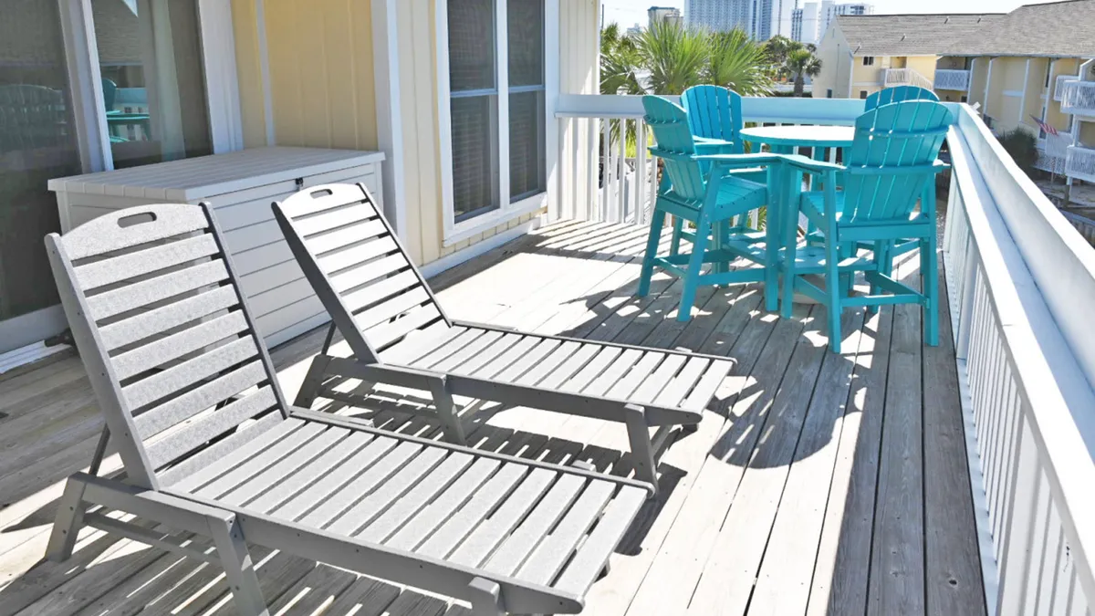 A private canal-front deck with two gray lounge chairs and a turquoise high-top table and chairs, all overlooking tranquil water and nearby buildings. The deck is bordered by a white railing, offering a peaceful spot for coffee or cocktails.