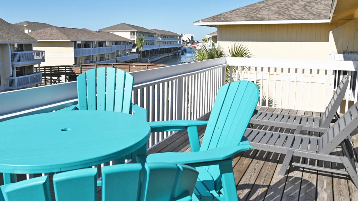 A private wooden deck overlooking the canal, featuring turquoise outdoor table and chairs and lounge seating. The view showcases boats in the canal and waterfront buildings, framed by a white railing—ideal for relaxing, dining, or enjoying cocktails at sunset.