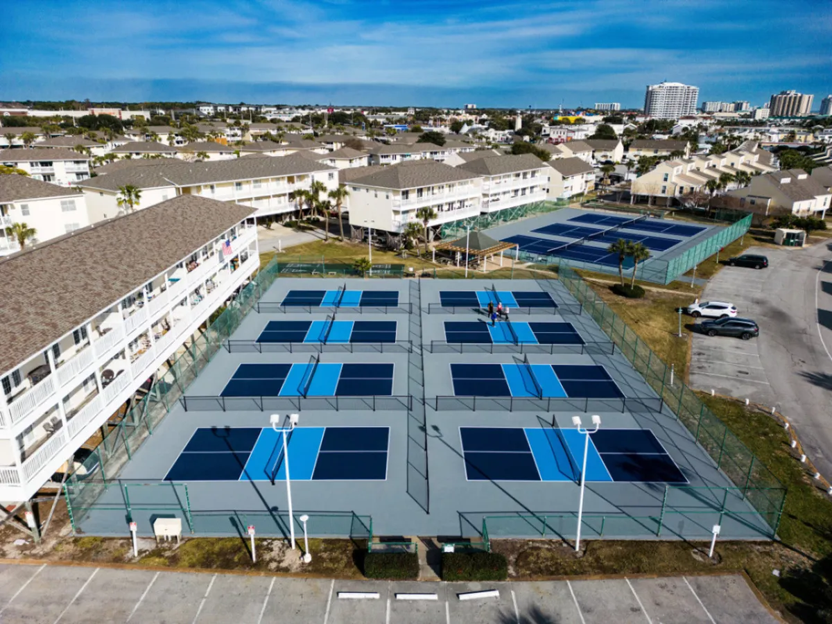 Multiple blue and gray pickleball courts sit adjacent to tennis courts, surrounded by resort buildings and a parking area. The courts are lively and inviting, designed for recreational play and social activity among guests and residents.