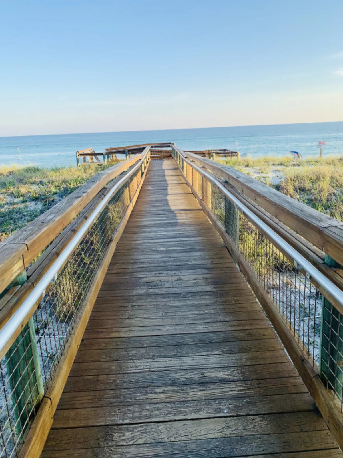 This exterior photo features a wooden boardwalk leading to the expansive sandy beach, bordered by coastal dunes and vegetation, with the calm blue ocean stretching to the horizon. It evokes the anticipation of reaching the beach and the natural beauty of the coastline.