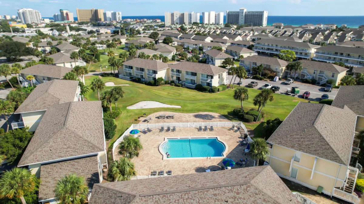 A central heated community pool is surrounded by lounge chairs and a green golf putting area. Rows of low-rise condos frame the pool, and in the distance, high-rise buildings and the ocean are visible, capturing the vibrant resort atmosphere.
