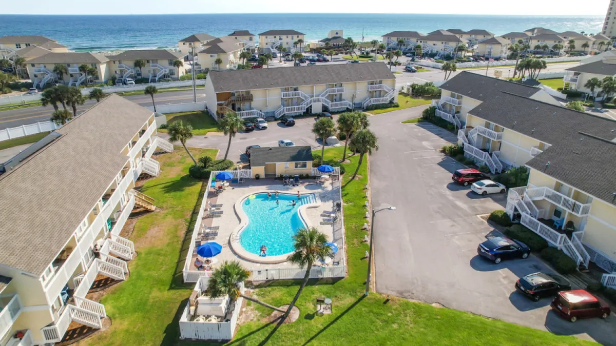 This exterior image showcases a vibrant central swimming pool with families and children enjoying the water. Surrounding the pool are two-story condo buildings with balconies, lush green lawns, and palm trees. In the background, the ocean and beach are visible, reinforcing the resort's proximity to the shoreline.