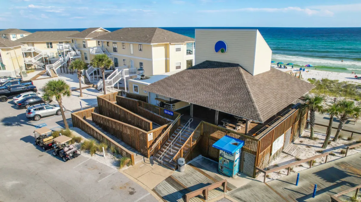 A wide exterior shot featuring the main resort building, a sandy beach lined with umbrellas, turquoise ocean waters, and a parking area with golf carts and cars. The lively beach scene suggests both adventure and relaxation, connecting the resort to the broader Destin beach experience.