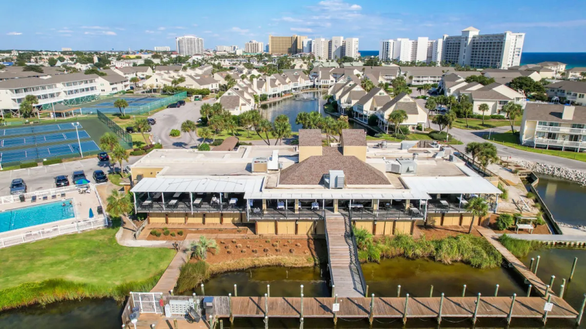 This exterior scene includes the central clubhouse with outdoor seating, adjacent tennis courts, a community pool, and canal-front docks. The image conveys a sense of an active, amenity-rich community with spaces for swimming, playing tennis, and relaxing by the water.