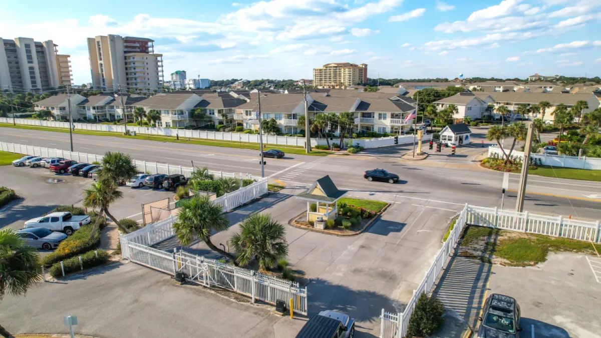 A secure, gated entrance with a guardhouse, palm trees, and landscaped greenery, with residential buildings visible in the distance. The road and entrance evoke the sense of arriving at an exclusive, well-maintained resort community.