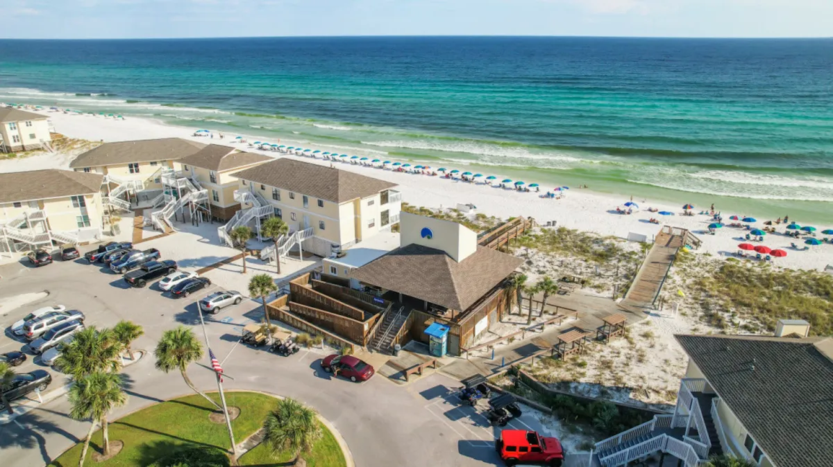 A vibrant white sandy beach lined with colorful umbrellas and beachgoers, with resort buildings and green ocean waves in the background. Pathways lead from the parking area to the beach, showcasing easy access to the shore.